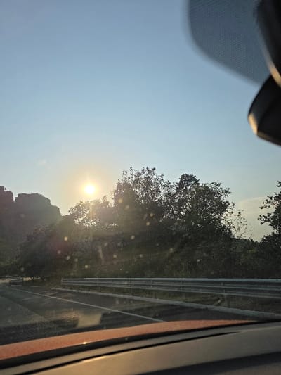 The image presents an outdoor scene viewed from the interior of a vehicle, located in the city of Tepoztlán, Mexico. The lower section of the frame features the vehicle's dark dashboard, detailed with a subtle reddish-brown trim, and a portion of the windshield exhibiting multiple circular light artifacts. A paved, two-lane road is visible in the midground, curving gently towards the left. Parallel to the right side of the road runs a three-tiered metallic guardrail. Beyond the road and guardrail, the environment consists of dense green foliage, primarily trees, which extend to the background. A distinct mountain or large hill is discernible on the far left. The sun, a bright, circular light source, is positioned low above the horizon on the left side of the frame, casting strong illumination across the landscape and causing direct lens flare. The sky overhead is clear and a uniform light blue. No individuals or animate subjects are visible within the frame.