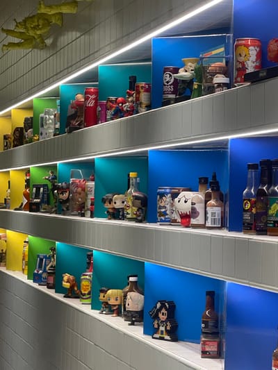 This is a well-lit, eye-level, medium shot of a display shelf featuring various collectible items and bottles. The shelves are angled, with each section illuminated by a colored light strip beneath it, creating distinct zones of yellow, green, and blue.
On the top shelf, a yellow, sculpted figure appears to be falling or in motion. Below this, the first visible section contains a red Coca-Cola can, a purple can with "BOSS COFFEE" text and a black silhouette of a man's head, a small Spiderman figurine, and other assorted figures and cans.
The next shelf down shows a variety of bottles and figures. Notably, there is a bottle with a yellow cap, a clear glass bottle, and several bottles of hot sauce with colorful labels. Figurines of characters from video games, such as Street Fighter, are prominently displayed.
Further down, more bottles and figurines are visible, including a Chupa Chups branded can, and characters that appear to be from anime or video games. The bottom shelf shows more hot sauce bottles and pixelated character figures.
The background consists of a light-colored, ridged wall. The overall impression is a curated collection of pop culture memorabilia and beverages. The scene does not provide any specific visual cues to pinpoint its location within Brisbane, Australia.