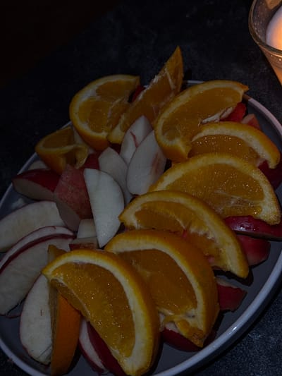 The image presents a close-up, high-angle view of a circular, light gray-rimmed plate filled with an assortment of sliced fruits, set within a dimly lit indoor environment, likely a private or commercial space in Bad Nauheim, Germany. The plate contains multiple wedge-shaped slices of oranges, distinguished by their vibrant orange pulp and visible peel, interspersed with slices of red apples showing white flesh, and segments of a lighter-fleshed fruit, consistent with peaches or nectarines, some exhibiting a faint pink hue near the skin. The fruit is densely arranged, covering the entire plate surface. Illumination, originating from the upper right, casts a soft glow on the fruit, and a portion of a glass object, possibly a candle holder with an internal light source, is visible in the upper right background. The surface supporting the plate is dark and appears to be textured, consistent with a countertop or table. No individuals are present.