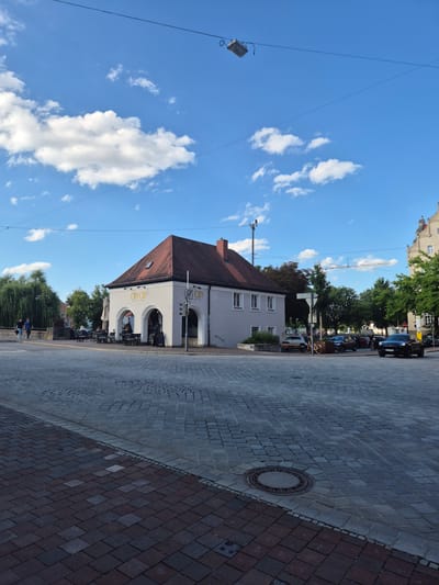 The image shows a street scene in Landshut, Germany. In the foreground, a cobblestone street is visible, with a manhole cover in the center. To the right, a black SUV is driving on the street. In the middle ground, a light-colored building with a red tile roof houses a cafe with outdoor seating. Several people are seated at tables outside the cafe, and others are walking in the background. A large tree with green foliage is on the left side of the building. The sky is blue with scattered white clouds. In the distance, there are more buildings and trees, and a construction crane is visible. The image appears to have been taken on a sunny day.