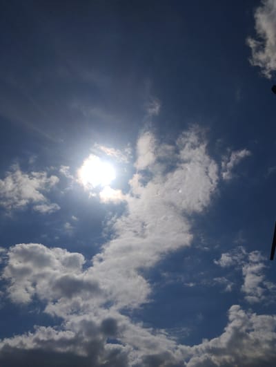 This image depicts a bright, sunlit sky with scattered clouds. The sun is prominently visible, shining through a patch of thinner clouds, creating a lens flare effect and casting visible rays of light. The sky is a deep blue, indicating it is daytime. The clouds vary in density, with some appearing wispy and others more substantial and puffy, casting subtle shadows. The overall impression is one of a clear, sunny day, likely late morning or early afternoon given the intensity of the sunlight and the presence of shadows. There are no visible people or objects in the sky, although a dark, thin vertical object is partially visible on the far right edge of the frame, suggesting it might be part of a structure or an antenna from ground level, or perhaps part of an aircraft. The setting is an open sky. There is no visible text in the image.