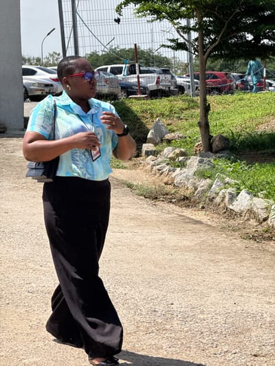 In Gwaska, Nigeria, on a bright day, a Black woman wearing red-rimmed glasses, a light blue and green patterned short-sleeved shirt, and a long black skirt walks on a paved path. She carries a black quilted purse on her left shoulder. Her right hand is raised, with her fingers extended as if gesturing or adjusting something near her chest. A lanyard with an ID card is visible around her neck. The path is lined with rocks and green grass on the right, and a concrete wall and a fence with vehicles in the background on the left. A small tree with dark green leaves stands between the path and the fence. The scene appears to be an outdoor area, possibly a parking lot or a facility.