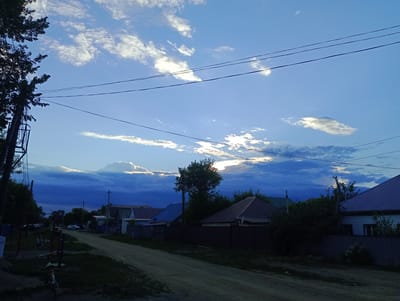 The image depicts a rural street scene in Atbasar, Kazakhstan, likely during the late afternoon or early evening, as indicated by the warm glow of sunlight breaking through dark clouds in the sky. The sky is a dramatic blend of deep blues and whites, with fluffy clouds catching the sunlight, creating a sense of impending twilight.
The foreground is dominated by a dirt road that winds its way into the distance. To the left, the silhouette of a large tree with its branches and leaves is visible against the bright sky, next to a utility pole with wires. Further down the road, there are a few houses with distinctive tiled roofs, characteristic of the region. Fences made of corrugated metal and wood line the properties.
In the middle ground, two people are seated near the tree on the left, appearing to be relaxing or conversing. A bicycle is parked in the foreground, suggesting a casual, everyday setting. A few cars are parked along the road and in driveways, indicating a residential area. The overall atmosphere is peaceful and serene, with a touch of natural drama from the sky. There is no visible text in the image.