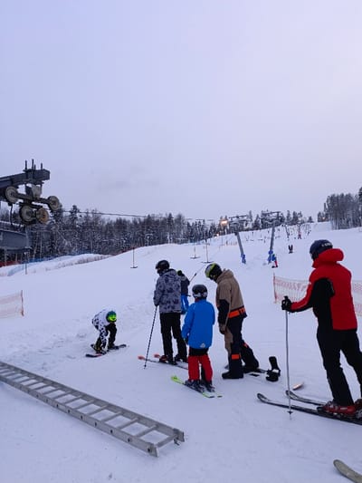The image captures a winter scene at a ski resort, likely during early morning, late afternoon, or on an overcast day, given the diffused light and the presence of artificial lighting on some ski lift structures in the distance. The ground is entirely covered in fresh, white snow, and a dense line of snow-laden evergreen trees forms the backdrop, separating the ski slope from the sky.
In the foreground, several individuals are gathered on the snow, equipped for skiing and snowboarding. To the far left, a person wearing a black and white checkered jacket and a yellow-green helmet is kneeling on a snowboard, appearing to adjust their bindings or prepare to stand up. Directly behind this person, on the left side of the image, a large, silver metal structure with multiple pulleys, part of a ski lift mechanism, rises against the sky. Below this, an orange safety net is visible along the edge of the slope. A long, silver-colored metal ladder lies horizontally on the snow in the bottom left corner of the image.
Moving from left to right in the foreground, there are four other individuals. The first, seen from the back, wears a grey and black patterned jacket and a black helmet, standing on skis and holding ski poles. Next to them, a child or young person in a bright blue jacket, red pants, and a black helmet is also on skis, facing away from the camera. Beside the child is an adult in a brown or khaki-colored jacket with orange accents, black pants, and a green-yellow helmet. This person is standing on skis, looking towards the left, with their back slightly turned to the camera. Furthest to the right in the foreground, another adult wears a vibrant red and black jacket, black pants, and a dark helmet, standing on skis and holding ski poles, also looking towards the left side of the frame. All individuals in the foreground are wearing helmets, indicating safety precautions.
Further up the snow-covered slope in the mid-ground and background, multiple ski lifts, possibly T-bar lifts, are visible, ascending the hill. Some of these structures have lights on them. Other skiers and snowboarders can be seen scattered across the wider slope, either heading up or preparing to descend. The overall activity suggests a busy day at a winter sports location, with people either waiting in line for lifts, taking a break, or preparing to engage in their chosen snow sport. No discernible text is visible in the image.