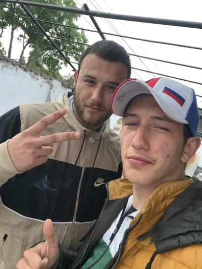 Two young men are posing for a selfie in what appears to be an outdoor location in Levski, Bulgaria. The man on the left is making a peace sign. The man on the right is wearing a cap with a Russian flag. The background includes trees, a fence, and a white building.