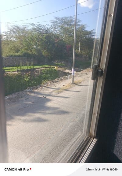 The image presents an outdoor scene viewed through a window, likely from a vehicle, with part of the window frame and glass visible in the foreground on the right side.

In the mid-ground, a light-colored, unpaved road or path extends from the center towards the upper right. Long shadows are cast across this path, suggesting strong sunlight. To the left of the path, there is a lower-lying area with vibrant green vegetation, possibly a cultivated garden or agricultural plot. Within this green area, several black containers or pots, arranged vertically, are visible in a row. Further back in this green section, a distinct bright yellow object, possibly a bucket or container, can be seen. Behind the green plot, there is a low stone wall.

Beyond the stone wall and green area, the background is dominated by numerous trees with varying foliage density. Some trees appear sparse, while others have denser green leaves, and hints of pink or red flowers are visible among some of the foliage, possibly bougainvillea.

A tall, slender utility pole stands prominently near the right edge of the road, extending upwards towards the sky. Some papers or posters are taped to its lower section. Overhead, power lines are visible against a clear, bright blue sky with a few wispy white clouds.

The window pane in the foreground shows reflections, particularly on the right side, subtly layering the view. The window frame appears to be a light-colored material, possibly metal or plastic, and a latch mechanism is visible on the right edge of the frame. To the immediate right of the window, a dark, textured surface, possibly the interior of the vehicle, is visible.

At the bottom left of the image, the text "CAMON 40 Pro" is printed, indicating the camera model used. At the bottom right, the camera settings "23mm f/1.8 1/418s ISO50" are displayed. The lighting suggests it is daytime, with clear weather conditions.