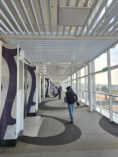 This is a medium shot, eye-level, wide-angle photograph of a modern airport jet bridge. The jet bridge is enclosed by large glass windows on the right, offering a view of an airport tarmac and terminal building. The ceiling is constructed with parallel white metal slats, and there are recessed lights and ventilation grilles visible. The walls on the left are white with large, dark purple abstract graphic patterns. The floor is carpeted with a patterned design of grey and dark grey curves. Several individuals are walking in the jet bridge. In the foreground, a man wearing a dark jacket, blue jeans, and black shoes is walking away from the camera, carrying a backpack and a small black bag. Further down the jet bridge, a person wearing a purple headscarf and carrying a tan bag is walking, followed by another person in a dark jacket and dark pants. The scene is brightly lit by natural light streaming through the windows. The location is identified as Ramunia Dua, Indonesia.
