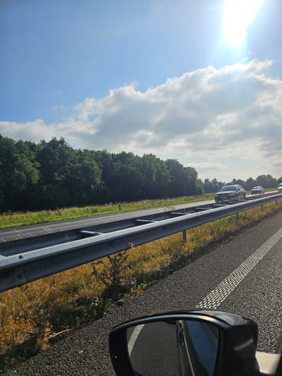 This media file captures a scene from a vehicle traveling on a highway, likely in Herwijnen, Netherlands, as indicated by the context. The perspective is from inside the vehicle, looking outwards and slightly downwards, with the side mirror of the car visible in the foreground on the right. The road surface is asphalt, marked with white dashed lines, indicating a clear lane. A metal guardrail runs along the side of the road, separating it from a grassy verge and then a dense line of trees.

The sky is a prominent feature, with a bright sun casting a glare on the left side of the image, suggesting it is daytime, possibly late morning or early afternoon given the intensity of the light. There are scattered clouds, some appearing fluffy and white against a clear blue sky. The sun's rays are strong enough to create lens flare.

Several vehicles are visible on the highway in the distance, moving in the same direction as the viewer's vehicle. These appear to be cars and possibly a van, following each other in a line. The trees bordering the highway are lush and green, indicating a likely warm season. The overall impression is of a clear, sunny day, with normal highway traffic. There is no visible text within the image. The scene evokes a sense of travel and the open road.