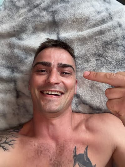 A man is lying on a gray, fuzzy blanket and smiling with his mouth open, revealing his teeth. He is pointing his index finger towards the viewer. He has dark hair, a tattoo on his chest, and another tattoo on his left shoulder. The image is a close-up of the man's face and upper body. The scene is indoors.