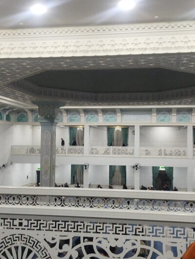 An interior view shows a multi-tiered mosque or religious building. Intricate white railings and pillars are visible in the foreground. A second level features arched openings with ornate carvings and green curtains. A third level echoes the second's design. People are scattered throughout the space, some standing on the upper levels. The ceiling is adorned with geometric patterns and recessed lighting. The overall impression is one of spaciousness and intricate design, typical of Islamic architecture.