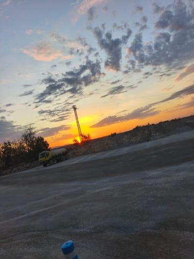 A yellow tanker truck is parked on a dirt road in what appears to be a construction or industrial area. The truck is facing left and has a large white cylindrical tank on its back. In the background, there is a power line tower with wires extending from it, and a sunset with orange and yellow hues is visible on the horizon. The sky is filled with scattered clouds. The foreground is a dusty, grey road surface. A blue and white striped traffic cone is visible in the lower right corner of the image. The scene is set in Riba-roja de Túria, Spain.