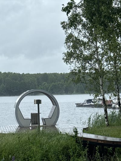 The image depicts a serene lakeside scene in Desnogorsk, Russia. In the foreground, lush green grass and reeds line the bank, leading to a wooden walkway. A distinctive circular bench with a central structure resembling a small table or receptacle is positioned on a wooden pier extending into the water. The lake itself is calm, with a distant treeline forming the background. Several boats are moored on the water, some appearing to be docked. The sky is overcast, suggesting a cloudy or potentially rainy day. The overall atmosphere is one of peaceful, natural beauty, with the man-made elements subtly integrated into the environment.