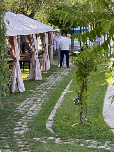The image depicts an outdoor seating area of a restaurant or cafe, likely located in Lozno, Bulgaria, given the context. The scene is dominated by a large, cream-colored canopy tent, supported by poles draped with beige fabric. Beneath the tent, several wooden tables and chairs are visible, suggesting a dining or lounging space.
A stone pathway winds through a grassy area, bordered by low stone edging. Several people are present. In the background, a group of four individuals are standing near a black metal fence, with a light-colored van visible behind them. One person in this group is wearing a light blue shirt, another a white t-shirt, and a third a light green shirt. A woman with blonde hair is also in this group. Another individual, a man in a white shirt and grey pants, is walking along the pathway. Inside the tent, a person with dark hair is seated at one of the tables.
The environment is verdant, with numerous trees and lush green grass. A small tree with a solar-powered lantern attached to its trunk is positioned near the pathway. The overall atmosphere suggests a pleasant, casual outdoor gathering.