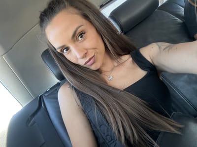 A woman with long, dark brown hair is pictured from the chest up, sitting in the back seat of a vehicle. She is wearing a black, sleeveless top and a silver necklace with a small, bejeweled dragonfly pendant. Her skin is light-toned, and she has brown eyes and dark eyebrows, with makeup applied. A tattoo is visible on her upper left arm. The interior of the vehicle appears to be dark leather. The image was taken in Opalchenets, Bulgaria.