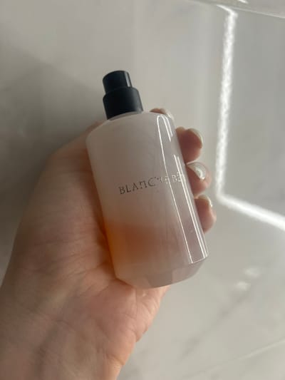 A hand with light-colored, manicured fingernails holds a white, cylindrical bottle with a black spray nozzle. The bottle has a pinkish hue towards the bottom. The text "BLANC le Bain" is embossed in a dark, stylized font on the side of the bottle. The background is a light-colored, textured surface, possibly a tiled wall, with a strong vertical highlight from a light source. The image does not provide any information about the location in the city of Нижние Муллы, Russia.