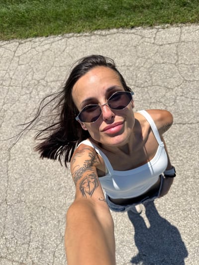 A high-angle selfie captures a young adult female on a cracked asphalt surface in Slavovitsa, Bulgaria. The subject, positioned centrally, has dark hair, partially windblown, and wears hexagonal-framed dark sunglasses. She exhibits a subtle smile with visible lips. Her attire consists of a white tank top and light-colored bottoms. Tattoos are visible on both arms: a detailed design featuring a stylized face split vertically on her left bicep, intricate floral patterns on her left upper arm, a small crescent-like symbol on her left forearm, and a minimalist sun outline on her right shoulder. A white-banded watch is present on her right wrist. Strong sunlight from the upper right casts distinct shadows, including the elongated shadow of her lower body and legs on the pavement. In the background, beyond the cracked pavement, is a strip of vibrant green grass.