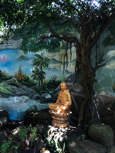 A golden Buddha statue is seated in meditation outdoors, positioned in the foreground of a mural. The mural depicts a landscape with pagodas, palm trees, and a body of water, all rendered in a vibrant, painted style. A large, gnarled tree with exposed roots dominates the right side of the frame, its branches extending over the Buddha statue and the mural. Lush green foliage from this tree creates a canopy, casting dappled shadows on the scene. The area around the statue and mural is comprised of large rocks and potted plants, suggesting a garden or shrine setting. The overall lighting is natural, with sunlight filtering through the leaves, creating a serene and tranquil atmosphere.