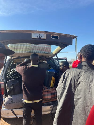 Four people stand near the open trunk of a car, packing it with luggage and other items. One person is reaching inside the trunk, while another is close by looking down into it. The car is filled with bags, a patterned blanket or cloth, and a yellow container. The sky is blue.