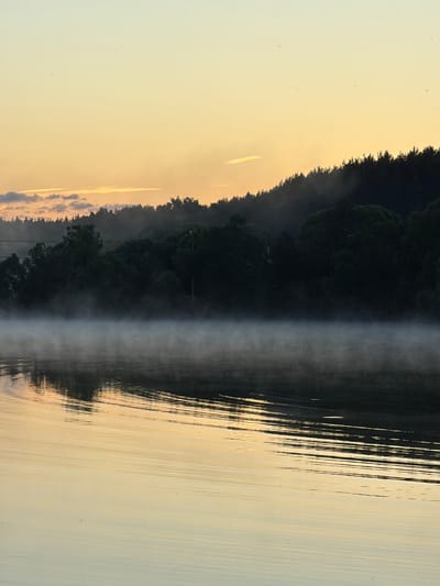 The image depicts a tranquil scene at the river in Satino-Russkoe, Russia, during sunrise or sunset. The sky is a soft gradient of yellow and pale orange, with wispy clouds visible near the horizon. A dense forest, silhouetted against the sky, forms the background, with some trees appearing slightly obscured by mist or fog. A thin layer of mist hovers just above the surface of the calm river, reflecting the warm tones of the sky. The water itself is smooth, with subtle ripples creating linear patterns across its surface. The overall impression is one of peace and natural beauty, characteristic of a serene morning or evening by the water.