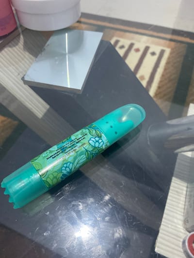 A teal-colored, double inhaler with a floral pattern is lying on a dark, reflective surface, possibly a table. The inhaler is capped with a clear teal plastic lid. Text is visible on the side of the inhaler, indicating it is a "Double Inhaler" with a Netto of 0.9 ml and 8 ml, and it is "Diproduksi oleh: PT. XYZ. Bekasi - Indonesia". The background shows a portion of a rug with brown and beige stripes, and a shiny rectangular object, possibly a metal plate, is visible to the upper left. The lighting suggests an indoor environment, with reflections indicating a light source. The scene appears to be a still life arrangement.