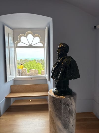 The image depicts an interior scene likely inside Pena Palace in Sintra, Portugal, based on the architectural style and views visible through the window. A bronze bust on a marble pedestal stands prominently in the foreground. The bust portrays a bearded man in ornate robes. Adjacent to the bust is a window with an ornate, decorative top. The window is open, revealing a scene outside featuring a person wearing a yellow shirt, standing at what appears to be a castle wall or parapet. Beyond the wall are green trees and possibly a glimpse of the ocean or a lake. A wooden bench is constructed to fit against the wall underneath the window, forming a seating area. The walls are painted a light blue-grey color, and the floor is made of light-colored wood panels. A security camera is discreetly mounted on the wall in the upper right corner.