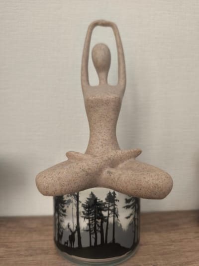 A beige, speckled sculpture of a stylized human figure in a meditative pose sits atop a cylindrical glass container decorated with a black and white forest scene featuring silhouettes of trees and deer. The sculpture's hands are raised above its head, forming a circle, and its legs are crossed in a lotus position. The glass container appears to be a candle holder or vase. The objects are placed on a wooden surface, with a textured, light-colored wall in the background. There is no indication of location within the city of Vyshhorod, Ukraine.