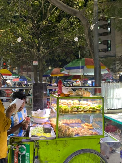 The image captures a nighttime street food market scene in Narayanganj, Bangladesh. A lime-green mobile food cart with wheels dominates the central foreground. The cart features a clear glass display case containing two shelves of prepared food items: the upper shelf holds multiple buns, some assembled as burgers with lettuce and patties, alongside various fried items; the lower shelf displays numerous breaded and fried snacks, consistent in size and shape.

On the cart's counter surface, a white rectangular tray contains shredded green vegetables and a yellow liquid, with a dark rectangular object situated beside them. A white plastic colander holds additional items. Behind the display case, a red plastic squeeze bottle, likely containing a condiment, stands upright. Partially visible to the left, an individual wearing a yellow garment holds a white paper wrapper. Behind the cart, a young male, appearing to be a vendor, wears a light blue and white striped Argentina football jersey and holds a red condiment dispenser, indicating active food preparation or service.

The background reveals a vibrant street environment illuminated by artificial lights. Multiple colorful market umbrellas in shades of red, yellow, blue, and green are visible, suggesting the presence of other food stalls. Numerous blurred figures populate the background, indicating a busy public space. A large tree with dense foliage occupies the upper portion of the frame, from which white cables and light fixtures are suspended. A utility pole or tree trunk is visible on the right, also with attached wiring. A multi-story building with illuminated windows can be seen in the distant background, confirming an urban setting. A green and white sign with Bengali text is affixed to the lower side of the food cart.