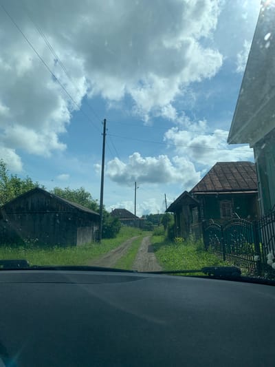The image shows a rural scene in Barsai, Russia. The perspective is from inside a vehicle, looking out through the windshield. The sky is a vibrant blue with scattered white, fluffy clouds, suggesting a sunny day, likely mid-morning or afternoon.
The foreground is dominated by the dark interior of the vehicle, with a textured dashboard on the right. A dirt road, lined with lush green grass and foliage, curves into the distance. Along the road, several wooden structures are visible. On the left side of the road, a tall wooden utility pole with wires stretches across the frame. Further down the road, a traditional-looking wooden house with a dark, gabled roof and a decorative window frame can be seen. Behind this house, a dark metal fence with an ornate design runs along the property. Another wooden outbuilding, possibly a shed or barn, with a weathered appearance, is located closer to the foreground on the left.
The overall impression is one of a peaceful, rural setting, possibly a village or a countryside road. The scene conveys a sense of calm and natural beauty. There is no visible text in the image.
