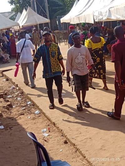 The scene depicts an outdoor gathering or market activity in Isinyi-Nando, Nigeria. In the immediate foreground, a blue plastic chair is partially visible on the left, next to a dirt ground area littered with plastic wrappers and small debris. This dirt area borders a slightly elevated, lighter-colored concrete pathway or curb running diagonally across the image.
Multiple individuals populate the midground. Two men are prominently walking towards the viewer: one in the center-left wears a black cap, a blue and yellow patterned short-sleeved shirt, black trousers, and dark sandals; the other in the center-right wears a light beige long-sleeved shirt with a cartoon hand graphic and "SHE MIN" text, dark shorts, and dark sandals. Behind the first man, another man walks away, wearing a white t-shirt with "Emanuell ONANOMA" printed on the back, light-colored trousers, and carrying a pink item in his left hand. To the far right, a man in a maroon shirt and dark trousers, facing away, is also walking. A woman in a yellow top and a patterned dark skirt is visible walking away from the viewer behind the central figures.
The background features numerous people congregated under several white canopy tents, suggesting a community event. A tall, dark metal structure, possibly a water tower or antenna, rises above the tents on the left side of the background. The environment is bright and sunny, indicated by distinct shadows cast by the individuals on the ground. A digital timestamp "19/12/2025 15:40" is present in the bottom right corner.