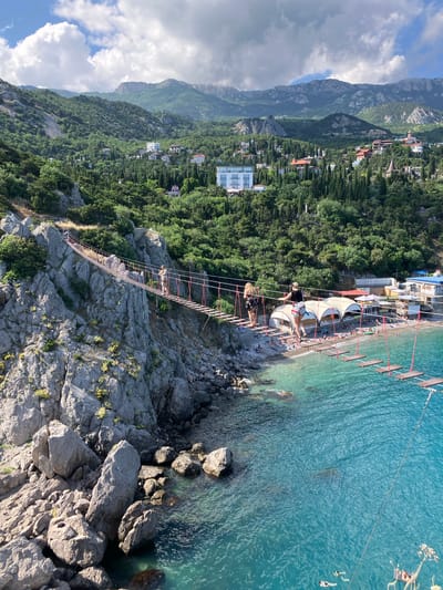 An outdoor, eye-level, medium shot shows a suspension bridge over a body of turquoise water. The bridge is made of wooden planks attached to ropes and extends from a rocky cliff face to a beach with buildings and awnings. Several people are on the bridge, walking in both directions. The cliff face is covered in rocks and vegetation. In the background, there are green hills with buildings scattered amongst the trees. The sky is cloudy. The scene is located in Simeiz, Ukraine, as indicated by the provided metadata.
