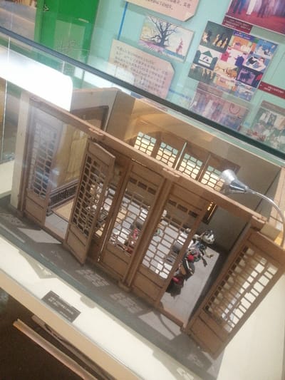 This image captures an indoor museum exhibit located in Fengtai District, China. The central subject is a detailed miniature architectural model, constructed from light-toned wood, depicting a traditional Chinese building structure. The model features multiple sets of lattice-patterned doors, several of which are open, revealing the interior. Upper sections of the model include windows with similar lattice designs.
Within the model's interior, several small, stylized figures are visible. One prominent figure, appearing to be a doll or puppet, features a white face, dark hair, and wears elaborate, multi-colored garments including red, blue, and black elements, positioned dynamically. Other less distinct figures or objects are also present inside the room. A small, silver-colored desk lamp on a flexible arm is positioned over the model, directing light into the scene. The floor within the model appears dark.
The model is displayed within a glass-enclosed case, indicated by reflections across the surface and edges of the glass. Beneath the model, a white exhibit label with black text, primarily in Chinese characters, is partially visible. The background consists of museum wall panels in shades of light blue-green and beige, displaying printed text in Chinese alongside various illustrative images or photographs. These include a depiction of a tree, multiple colorful cartoon-like scenes, and what appear to be historical photographs or artworks. A large, green rectangular object is partially visible on the far left of the background. The photograph is taken from an elevated, oblique perspective, looking down into the exhibit.