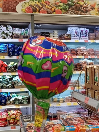 This media file is a photograph taken inside a brightly lit supermarket in Pavlovsky Posad, Russia. The primary subject is a large, vibrantly colored lollipop wrapped in shiny plastic, held in the foreground, obscuring a significant portion of the shelves behind it. The lollipop has a playful design with cartoonish characters and a pattern of stripes in shades of pink, blue, and green.

The background reveals rows of refrigerated shelves stocked with various food products. On the top shelf, a display of what appears to be meat and salad is visible. Below that, shelves are filled with packaged goods such as bags of chips or snacks, cartons of sausages, and smaller containers of food items. Some text is visible on packaging, including "Ricco", "ХЕЛЯМАНИС", and "11 Микоян".

The overall impression is a typical grocery store environment, likely during daytime hours given the bright artificial lighting. There are no people prominently featured in the image. The focus on the lollipop suggests it might be a promotional item or a product being showcased. The scene is focused and does not provide strong emotional cues. The presence of Russian text on the packaging confirms the location context.