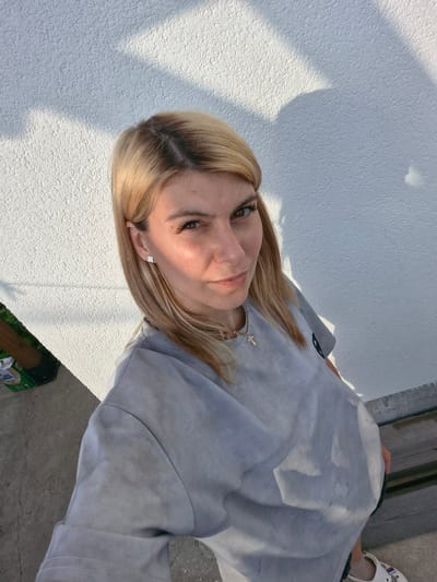 This is a selfie taken by a young woman with blonde hair. She is wearing a light gray t-shirt and a necklace with a cross pendant. She has a small clover-shaped earring in her left ear. She is standing in an outdoor setting, with a white textured wall behind her. The sun is shining, casting shadows on the wall, suggesting it is daytime. The ground appears to be concrete or pavement. In the background, a green box with some text is visible. The woman is smiling gently, and her eyes are looking towards the camera. The overall mood appears casual and relaxed.