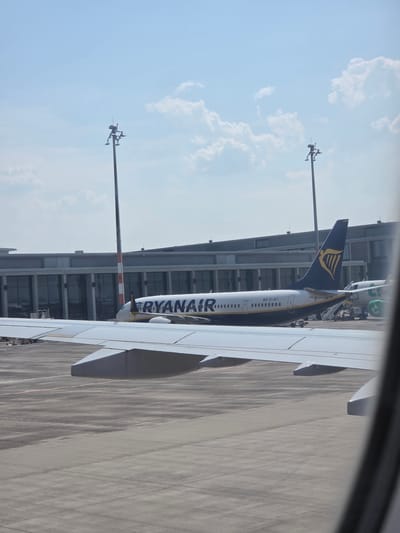 In Schönefeld, Germany, a Ryanair airplane is parked at an airport gate. The airplane, with the registration EI-IKT, is white with navy blue accents and features the distinctive Ryanair logo on its tail. The aircraft's wing is visible in the foreground, and the terminal building with its large glass windows is in the background. Several airport lighting poles are also present on the tarmac. The sky is a clear blue with scattered white clouds. The scene captures a typical airport environment on a bright day.