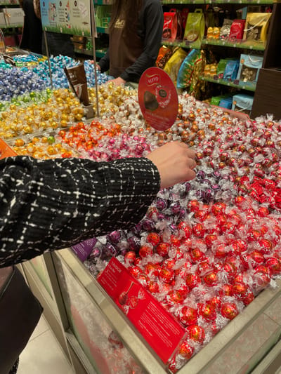 The scene depicts an interior view of a Lindt chocolate store or concession located in Marratxí, Spain. In the foreground, the left arm of an individual, wearing a black and white patterned tweed-style jacket, extends into a large, multi-compartment display bin filled with bulk, individually wrapped Lindor chocolates. The hand is positioned above sections containing red and purple foil-wrapped truffles.
The extensive display bins showcase a spectrum of Lindor chocolate varieties, color-coded by wrapper, including blue, green, yellow, orange, pink, purple, red, and silver/white. A red oval-shaped sign, centrally placed above the bins, reads "NUEVO" (New) and identifies "LINDOR Corazón 70%." A rectangular red sign at the bottom right of the display labels a section "LINDOR Leche" (Lindor Milk). A brown paper scoop or bag holder is visible within one of the central bins.
In the background, a store employee, partially visible, stands behind a counter. This individual wears a brown apron featuring the Lindt logo. Behind the employee, shelving units are stocked with various packaged chocolate products, gift bags, and boxes, indicative of a retail setting. The area is brightly illuminated, and the flooring appears to be light-colored tiled material.