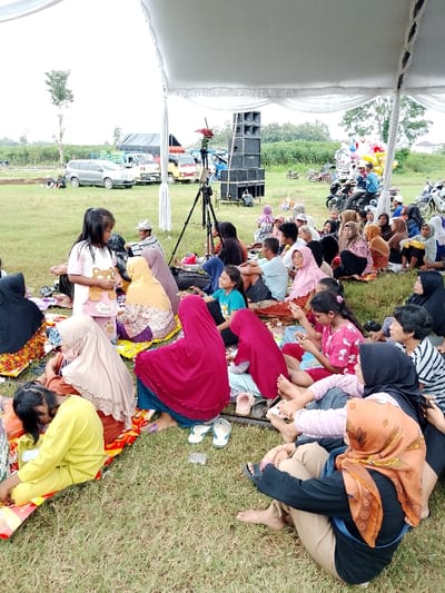 The scene takes place outdoors under a white tent, on a grassy area, in the city of Kecamatan Margorejo, Indonesia. The coordinates are -6.735 latitude and 111.016 longitude. There are approximately 30 people seated on the ground in a semi-circle facing inwards, seemingly attending an event. Most are wearing casual clothing, some are wearing headscarves. There are several groups of people: two adults and a young girl are seated in the foreground; three adults are seated further back; a group of 11 adults and children are seated in a row near the center of the image; a group of 5 adults are seated near the edge of the image; and a large group of 9 adults are seated in the background. The scene contains numerous personal belongings including handbags, water bottles, and food items. Several adults are engaged in conversation. There is a large speaker system to the left of the image. There are several parked vehicles in the background, including a grey car and several trucks.
