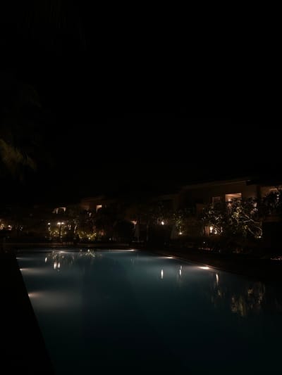 The image captures a serene nighttime scene at what appears to be a resort or hotel property in Betalbatim, India. A large swimming pool dominates the right side of the frame, its calm, dark blue surface reflecting multiple bright, elongated streaks of light from unseen sources. These reflections suggest the presence of underwater or edge lighting, as well as ambient illumination from the surroundings.

Along the left edge of the pool, several multi-story buildings are visible, their architecture hinting at accommodation or facility structures. Numerous windows and balconies on these buildings are softly illuminated, adding to the warm glow of the scene. Lush trees and dense shrubbery are interspersed among the buildings and line the pool's edge, some parts of the foliage also illuminated by lights, creating a tropical and inviting atmosphere. The sky is completely dark, indicating late evening or deep night. The foreground and the far left of the image are very dark, suggesting an expansive unlit area or deep shadow.

No people are visible, contributing to a quiet and peaceful ambiance. The still water and consistent lighting convey a sense of tranquility and relaxation. There is no visible text or identifiable activity taking place, rather the image focuses on the calm beauty of the illuminated architectural and natural elements at night.