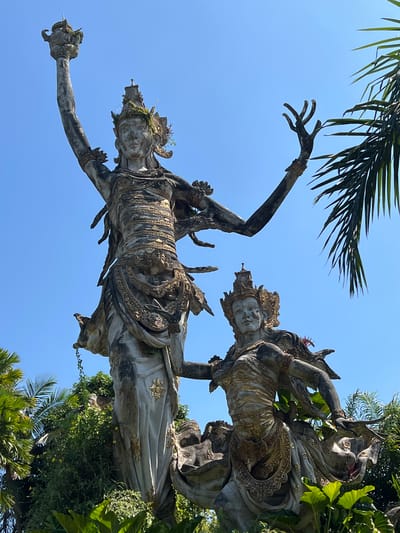 The image captures a view of a Balinese sculpture in Kedewatan, Indonesia, against a clear blue sky. The sculpture depicts two human-like figures adorned with intricate detailing and crowns. The statues are positioned amongst lush foliage, with palm fronds visible both above and on the right side of the image. The statues are a grey, stone-like material with gold detailing. The overall orientation of the image suggests it was taken from below, looking upwards towards the statues and the sky.