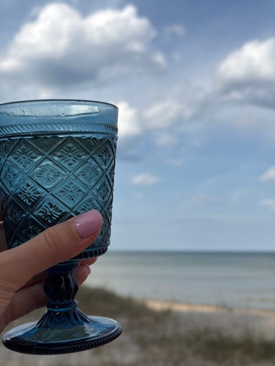 A person holds a blue decorative glass in front of a blurred background of a cloudy sky and a light-colored landscape, possibly a beach or field, in what appears to be the city of Plieņciems, Latvia. The glass has a geometric pattern on the body and a goblet-like shape with a flared base. The person's hand, with a light pink nail polish, is partially visible.