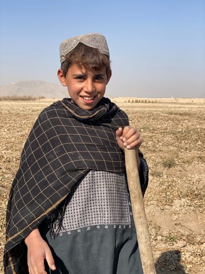 A young male subject, appearing to be a boy, is depicted standing in an open, arid field in Mandisar, Afghanistan, under a clear, light blue sky with distant haze. He has dark brown, curly hair and is smiling directly at the viewer, revealing teeth. He wears a light grey patterned cap, a dark grey or black shalwar kameez with a white and black geometric patterned bodice, and a black shawl draped over his shoulders featuring a grid pattern of thin light-colored lines. His right hand grasps a weathered, light-colored wooden stick, which rests vertically beside him. The ground consists of dry, light brown soil with sparse vegetation and scattered small rocks. In the background, low, distant mountains are visible on the left, while a long, light-colored wall or structure extends horizontally across the mid-ground to the right, with a faint white vertical structure visible further beyond it.