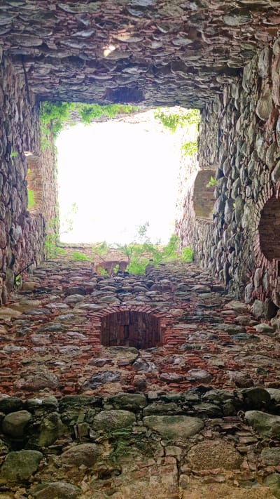 The image shows the interior of a ruined stone structure, likely a castle or fortification, looking upwards towards an open sky. The walls are constructed from rough-hewn stones and bricks, some of which are covered in moss and vegetation. There are arched openings visible in the walls, and the floor appears to be made of rubble. The scene is characterized by a sense of age and decay. This is a view of the ruins of the Embūte castle mound and manor house, located in Embūte, Latvia.