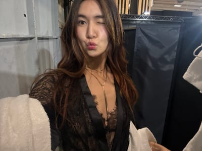 An adult female is depicted in a close-up, chest-up selfie taken in Issy-les-Moulineaux, France. She possesses long, wavy brown hair with lighter highlights, a light complexion, and is looking directly at the camera with her left eye winked shut and lips puckered into a "kiss" gesture. She is wearing a black lace bralette or camisole beneath a partially open black satin robe or wrap with lace trim. Two gold-toned necklaces, a choker and a longer lariat-style pendant, adorn her neck. Her right hand, partially visible on the lower right, holds the camera and clutches a white, fluffy towel. Another white towel is draped over her left arm. The background to the left consists of a light grey, paneled wall. To the right, a dark, possibly black fabric partition hangs from an overhead metal truss structure. Exposed ceiling elements, including wooden slats and metal beams, are visible above the partition. Another white towel is partially visible on the far right, hanging in the background.