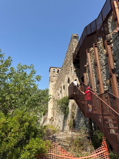 An exterior, low-angle shot shows a weathered stone structure resembling a castle or ancient ruin, with a modern, rust-colored metal staircase and walkway attached. The sky is a clear, bright blue. To the left, a vibrant green tree with dense foliage partially obscures the stone building. The stone structure has a tower-like section with arched openings. The metal staircase, made of grated material, winds its way up the side of the stone building, supported by vertical steel beams. Two people are visible on the staircase: a person in a white shirt and dark pants is higher up and further away, while a child wearing a red cap and shorts is closer to the foreground, ascending the stairs. The environment appears to be a historical site with some areas cordoned off by orange construction netting in the foreground. The scene suggests a warm, sunny day. The location is the Doria Castle in Dolceacqua, Italy.