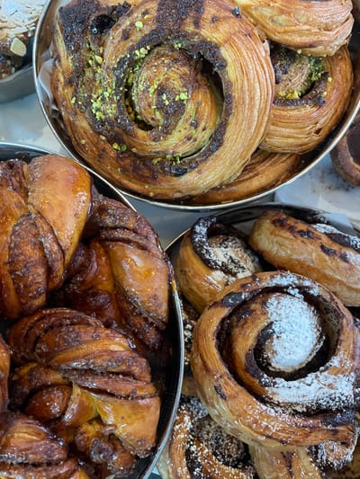 Several metallic containers hold assorted pastries. One container features spiral-shaped, flaky pastries, some sprinkled with pistachio. Another container presents similarly shaped pastries, dusted with powdered sugar and filled with chocolate. The last container contains glazed, knotted sweet rolls with a rich, brown color. The pastries appear fresh and appealing.