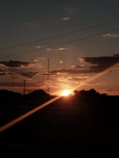 This is a vertical, medium-shot photograph of a sunset in Aqadyr, Kazakhstan. The sun, a bright, glowing orb, is low on the horizon, casting a warm, orange light across the sky and through the clouds. The sky transitions from a deep twilight blue at the top to shades of orange and pink near the horizon. Scattered, dark clouds are silhouetted against the colorful sky, some illuminated by the setting sun.

In the foreground, the landscape is mostly silhouetted against the bright sky. There are hills and trees on the horizon, as well as two tall radio towers, one on the left and a taller one in the center. A bright, diagonal light streak, likely a lens flare, cuts across the lower right portion of the frame, originating from the setting sun. Two thin, dark horizontal lines are visible across the upper half of the image, suggesting overhead wires. The overall mood of the image is serene and peaceful, capturing the natural beauty of a sunset in a rural or semi-rural setting. The time of day is clearly sunset.
