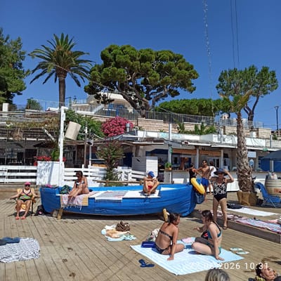 This image depicts a lively outdoor scene at a beachside establishment in Trani, Italy, on June 22, 2026, at 10:31 AM. Several individuals are enjoying a sunny day, with some relaxing on the wooden deck and others near a blue boat.
In the foreground, two women are seated on blankets on the wooden deck. One woman, wearing a black and white patterned swimsuit and sunglasses, is holding her hat. Another woman, in a teal bikini, is sitting cross-legged and gesturing with her hand. To the left, a woman in a colorful swimsuit and pink cap is seated in a beach chair, facing left.
A distinctive blue boat, serving as a seating area or prop, is positioned on the deck. A young man in red shorts and a blue cap is sitting on the edge of this boat. Another woman in a black and white swimsuit is sitting on the boat's edge, her legs extended. A yellow inflatable ring is visible near the boat.
In the background, a row of buildings appears to house a bar or restaurant with outdoor seating. People are visible standing and interacting within this area. Palm trees and lush green trees provide a tropical ambiance, contrasting with the architectural elements. The sky is clear and blue. The wooden deck is sparsely populated with beach towels, bags, and flip-flops. The overall atmosphere suggests a relaxed, recreational setting.