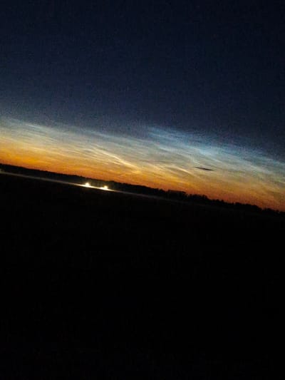 The image captures a nocturnal landscape in Kyviškės, Lithuania, featuring a gradient of sky colors from deep indigo at the top to warm orange and pale blue near the horizon. Wispy, luminous clouds, possibly noctilucent clouds, are illuminated by an unseen light source, creating an ethereal effect across the sky. Below the sky, a dark, silhouetted treeline marks the transition to the ground. In the foreground, a flat, dark expanse, likely a field, dominates the lower half of the image. A faint line of lights, possibly from vehicles or distant buildings, is visible along the horizon, suggesting human presence in the area. The overall scene is serene and atmospheric, characterized by the contrast between the dark earth and the brightly lit, high-altitude clouds.