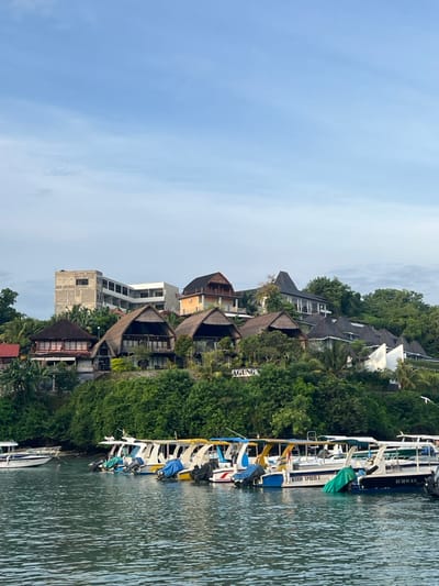 This image presents a coastal scene in Toyapakeh, Indonesia, under a clear, light blue sky with sparse, high-altitude clouds. The foreground consists of calm, dark green-blue water, occupying the lower third of the frame. Eight passenger boats, predominantly white with yellow, blue, and green accents, are moored in a line across the water, facing towards the right. These vessels are equipped with outboard motors; specific names "KARD I XPRESS 3" and "ICHWAN" are visible on two boats. Several boats feature blue or green tarpaulins covering sections. A smaller, white boat is partially visible on the far left.
The midground and background feature a steep, lush green hillside densely covered with tropical foliage. Numerous buildings are constructed into this terrain. A multi-story concrete structure, appearing to be under construction or unfinished, is located on the upper left portion of the hillside. Below and to the right, several buildings exhibit prominent thatched roofs, characteristic of traditional Balinese architecture. Other structures with dark, conventional roofs and modern designs are interspersed among the thatched-roof buildings. A white sign, partially obscured by greenery, displays the text "AGUNG" on the hillside. No individuals are visible in the scene.