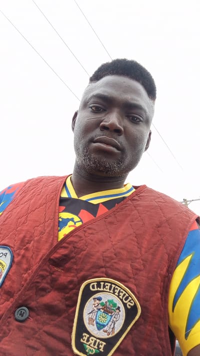 A medium-shot, low-angle image shows a dark-skinned man with a close-cropped haircut and a patchy beard wearing a maroon quilted vest over a colorful shirt. The vest has two patches, one partially visible on the upper left and the other fully visible on the lower right. The full patch reads "SUFFIELD FIRE CT". The man's face appears moist. Above him, against a white sky, are several parallel power lines. Given that Kawo is in Nigeria, and the patch on the vest reads "SUFFIELD FIRE CT," it is unlikely the photograph was taken in Kawo. The environment appears to be outdoors under overcast lighting.