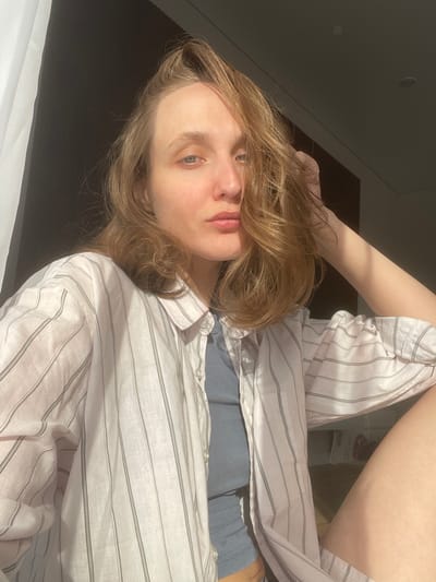 A woman lies on a dark surface, possibly a bed or floor, in an interior space. She is facing the camera with her head tilted. She wears a striped, light-colored, long-sleeved shirt over a blue garment. Her hair is blonde and wavy, partially obscuring her face. The lighting is uneven, with strong shadows cast on the left side of the frame and brighter illumination on the right. The background is blurred, but suggests a room with varying levels of light and dark. There is no information about the location in the city of Sepych, Russia.